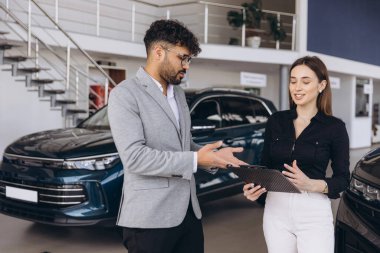 Car salesman explaining features of new car to female customer holding a clipboard