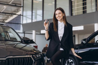 Smiling saleswoman presenting a car key while seated on a new vehicle in a vibrant dealership showroom, radiating confidence and success