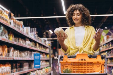 Customer comparing products on a smartphone while grocery shopping in a supermarket, making informed choices about healthy options