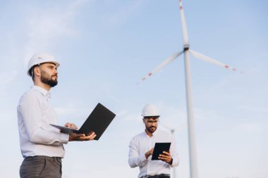 Two male engineers using laptop and tablet working next to wind turbines for sustainable energy production under blue sky