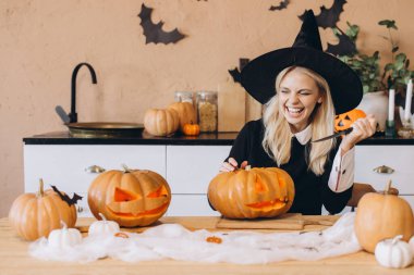 Young woman laughing and wearing a witch hat while joyfully carving pumpkins in the kitchen for a festive Halloween party celebration