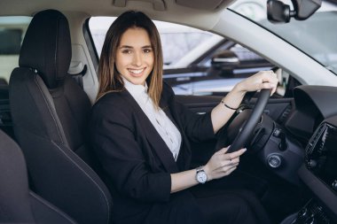 Young saleswoman sitting inside new car and holding steering wheel, smiling at camera in car dealership showroom