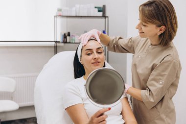 Beautician wearing gloves, gently touching the forehead of a patient holding a mirror in a serene beauty clinic setting