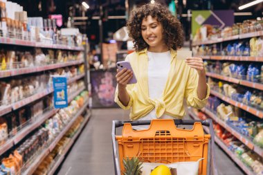 Smiling curly woman using smartphone and shopping list while pushing shopping cart in supermarket