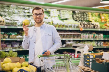 Customer selecting groceries in supermarket, holding apple and shopping list, surrounded by fresh fruits and vegetables