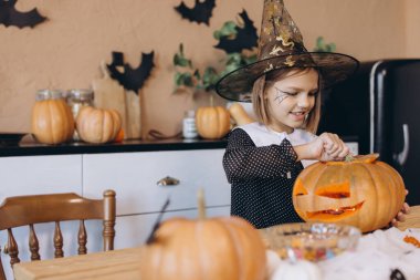 Smiling girl wearing a witch hat, happily carving a Halloween pumpkin while sitting at a wooden table in a cozy kitchen