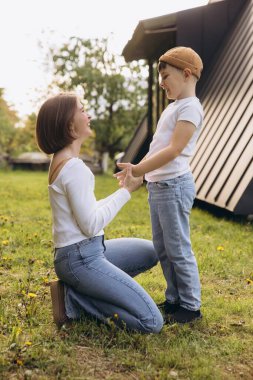 Happy mother kneeling on grass holding her son's hands, smiling and playing together in front of their modern house, enjoying a sunny day outdoors