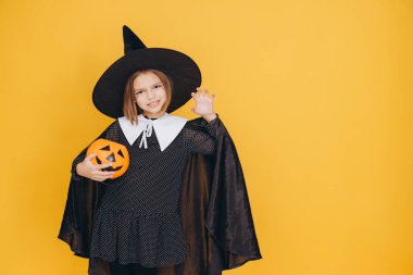 Little witch holding a pumpkin basket with painted face, wearing black and white dress and hat, posing on a vibrant yellow background for Halloween