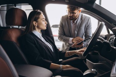 Salesman explaining car features to businesswoman sitting in driver's seat at dealership