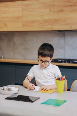 Focused pupil writing in notebook, sitting at table with tablet, headphones and colored pencils, studying at home in bright kitchen