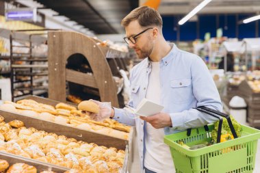 Man wearing transparent gloves choosing bread from bakery section in supermarket, holding shopping basket and reading shopping list