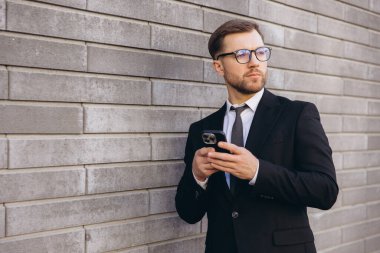 Modern businessman using smartphone standing next to a brick wall, looking away