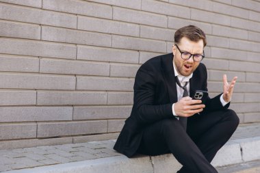 Frustrated businessman experiencing problems with mobile phone while sitting on the curb