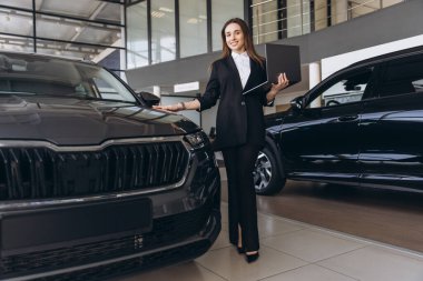 Saleswoman presenting a new car to a customer while holding a laptop, engaging in a modern car dealership environment