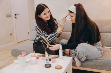 Two teenage girls sitting together on a cozy sofa, enjoying makeup application while sharing laughter and creating lasting memories