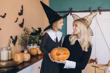 Mother and daughter smiling and looking at each other, wearing witch costumes and holding a carved pumpkin in their hands, preparing for Halloween