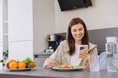 Young woman enjoying a nutritious meal while browsing her smartphone in a stylish, modern kitchen, embracing a healthy lifestyle and wellness