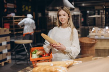 Young woman selecting a fresh baguette while shopping for groceries in the bakery section of a bustling supermarket