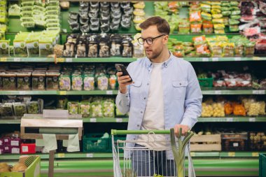 Man using a smartphone while pushing a shopping cart in a supermarket, choosing fresh produce