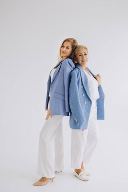 Studio portrait of elegant mother and daughter businesswomen posing back to back, wearing blue jackets and white pants, on white background