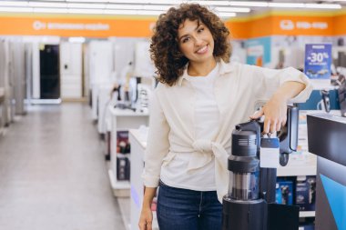 Smiling woman selecting a new cordless vacuum cleaner in an appliance store, enjoying shopping for household goods