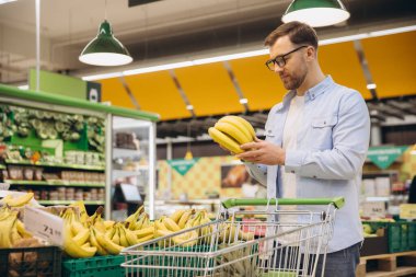 Customer selecting fresh bananas in grocery store, holding a bunch and pushing a shopping cart with more bananas inside