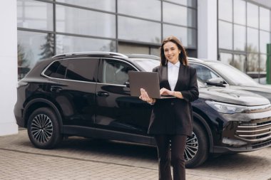 Saleswoman holding laptop standing in front of new black cars at car dealership, automotive industry