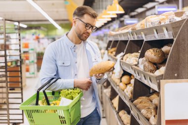 Customer choosing bread from shelf in supermarket bakery section holding shopping basket with groceries