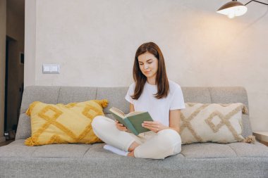 Young woman enjoying a quiet moment at home, comfortably seated on her sofa and engrossed in reading a book