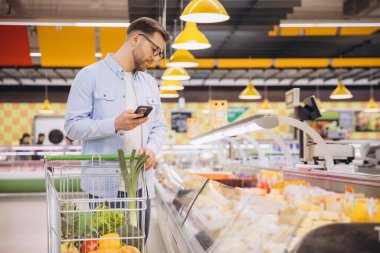 Man checking shopping list on his phone while pushing a cart full of vegetables and fruits in a supermarket