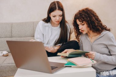 Two focused teenage girls using laptop and books, learning together at home, preparing for exams or doing homework