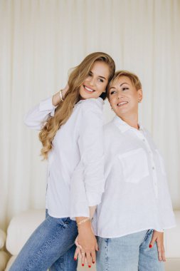 Happy mother and daughter holding hands, wearing white shirts and jeans, standing in a bright interior, enjoying a tender moment of family bonding