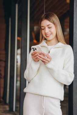 Smiling woman using smartphone while leaning against a wall, enjoying mobile technology and online communication
