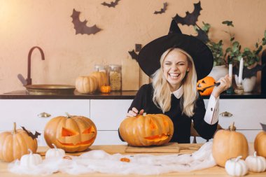 Happy witch laughing and joyfully carving pumpkins in the kitchen, preparing for a festive Halloween party filled with excitement and cheer