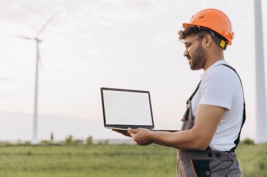 Indian engineer wearing an orange helmet and overalls is holding a laptop with blank screen in a field with wind turbines