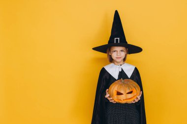 Young girl wearing witch costume holding carved pumpkin posing on yellow background for Halloween party