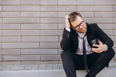 Stressed businessman scratching head while looking at his smartphone sitting on sidewalk