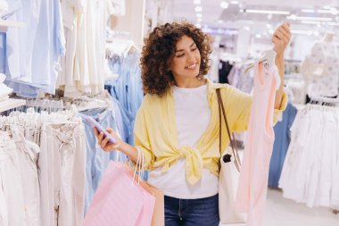 Happy customer holding pink shopping bags and using mobile phone while choosing clothes in fashion store