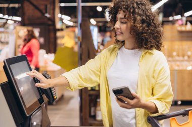 Woman using self service checkout kiosk in a modern supermarket, efficiently paying for groceries with a smartphone and enjoying a seamless shopping experience