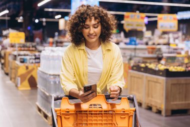 Smiling woman using a shopping list app on her smartphone while pushing a cart through the grocery store, selecting fresh products