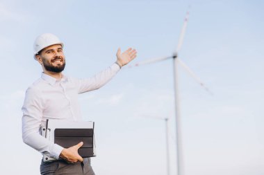Smiling engineer holding a laptop while showcasing wind turbines at a sustainable energy farm, highlighting the importance of renewable energy