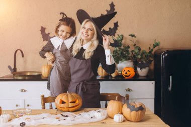 Mother wearing a witch hat and daughter joyfully carving pumpkins together at home, preparing for a festive Halloween party