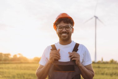 Smiling Arab engineer standing confidently in front of a wind turbine during sunset, embodying pride in sustainable energy innovation