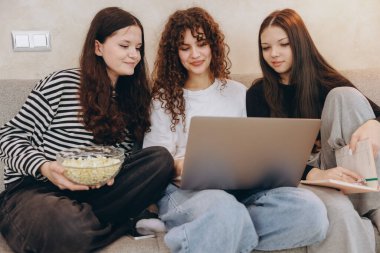 Three teen girls are sitting on a couch, watching a movie on a laptop and eating popcorn from a bowl