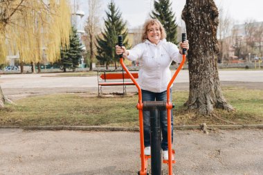 Elderly woman exercising on elliptical cross trainer in a public park, enjoying healthy lifestyle and outdoor activity