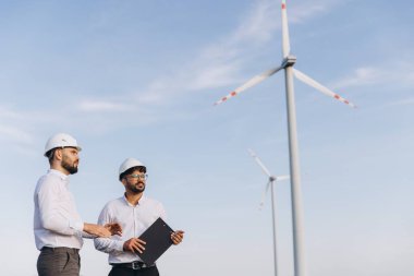 Two engineers wearing helmets are discussing about a wind turbine farm for sustainable energy production