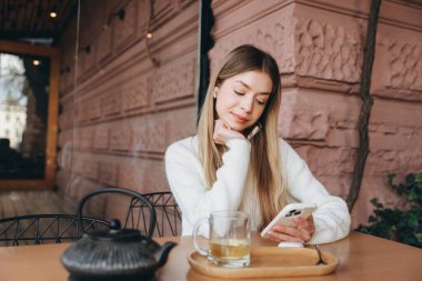 Happy freelancer working remotely from outdoor cafe, using mobile phone and drinking herbal tea