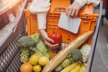 Customer holding a red bell pepper and checking her shopping list while buying fresh produce at the supermarket