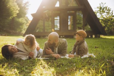 Happy family lying on grass enjoying sunset in front of their modern a frame cabin in the woods