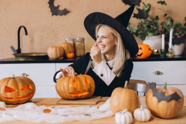 Happy woman wearing a witch hat, joyfully carving pumpkins for a festive Halloween party at home, surrounded by autumn decorations